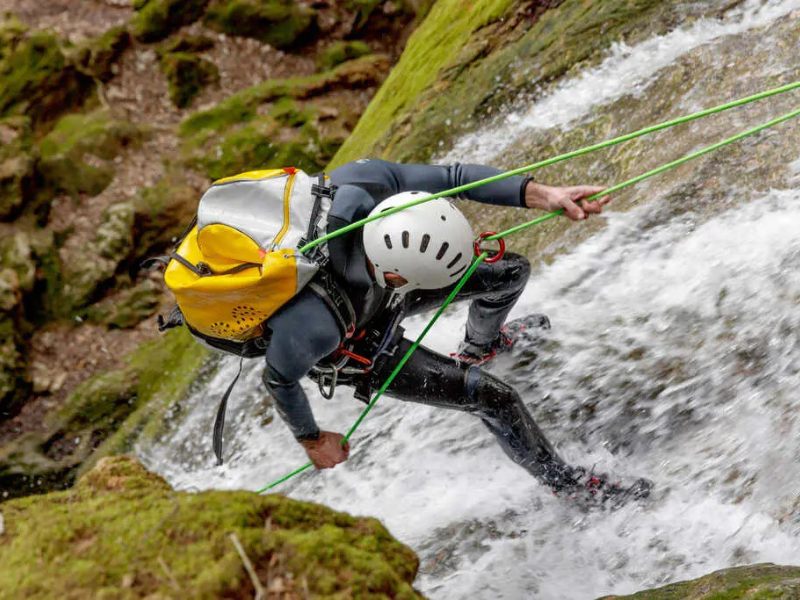 Um alpinista com capacete branco desce de rapel uma cachoeira molhada carregando uma mochila amarela.