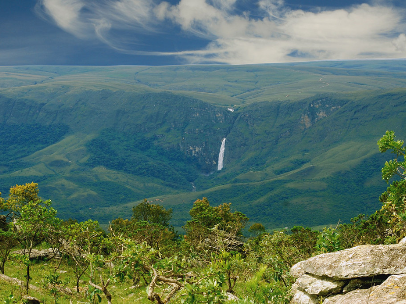 Montanhas verdejantes com uma cachoeira ao longe sob um céu nublado, vistas de um mirante rochoso.