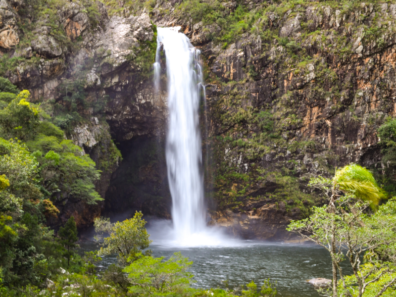 Alta cachoeira despencando em uma piscina em meio a penhascos rochosos e folhagem verdejante.