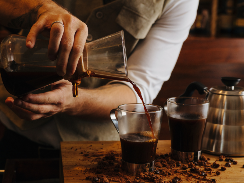 O barista serve café de uma garrafa térmica de vidro para uma xícara transparente ao lado de uma chaleira de metal em um balcão de madeira.