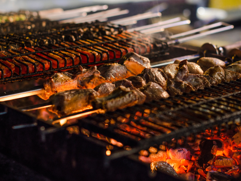 Carnes variadas grelhando em uma churrasqueira a carvão, com brasas incandescentes e espetos de metal.