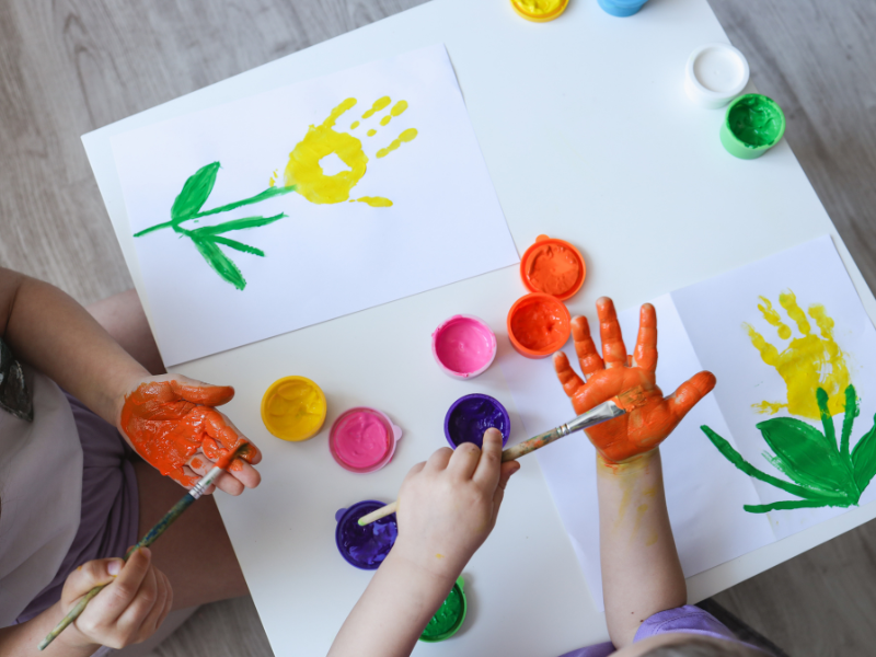 Crianças pintando impressões de mãos coloridas em papel, sentadas em uma mesa com potes de tinta e pincéis.