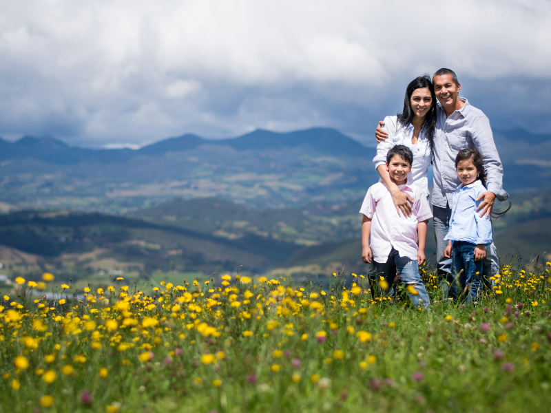 Família em pé num prado florido com montanhas ao fundo.