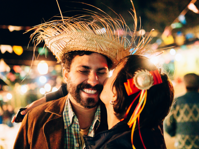 Duas pessoas sorrindo e dançando em uma festa animada ao ar livre; uma delas usa um chapéu de palha, com luzes de cordão ao fundo.