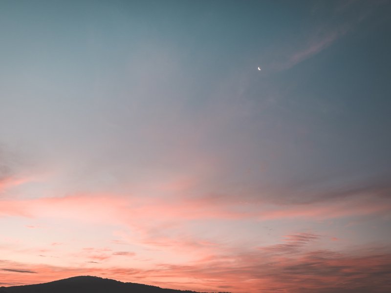 Céu rosa e azul ao pôr do sol sobre colinas escuras, com nuvens finas e uma lua crescente brilhante.