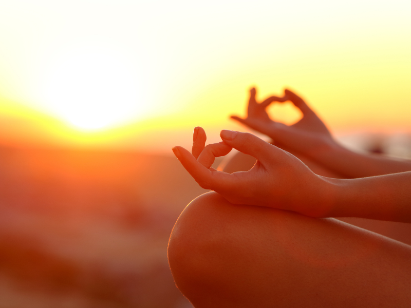 Mãos em posição de meditação ao pôr do sol, silhuetadas contra um céu alaranjado brilhante.