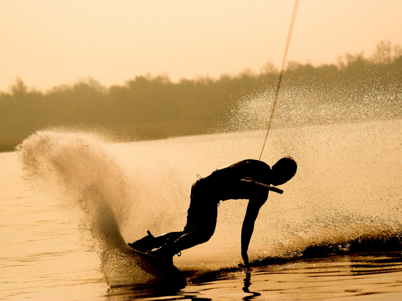 Silhueta de um praticante de wakeboard deslizando sobre a água ao pôr do sol, lançando um grande arco de água.