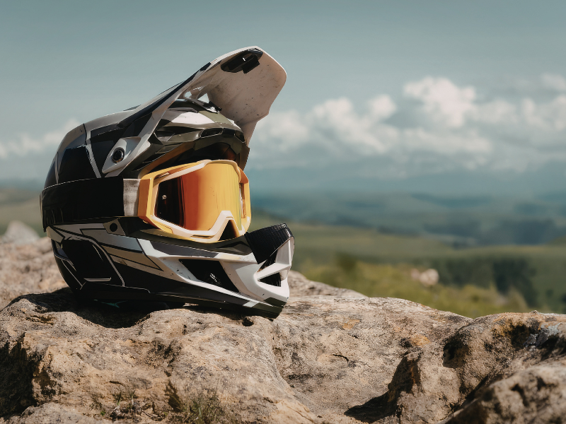 Capacete preto de trilha com óculos âmbar apoiado em uma rocha, com montanhas e céu ao fundo.