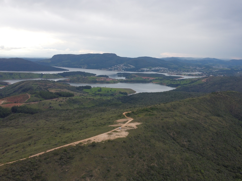 Ampla vista aérea de colinas verdejantes, uma estrada de terra sinuosa e um lago sob um céu nublado.