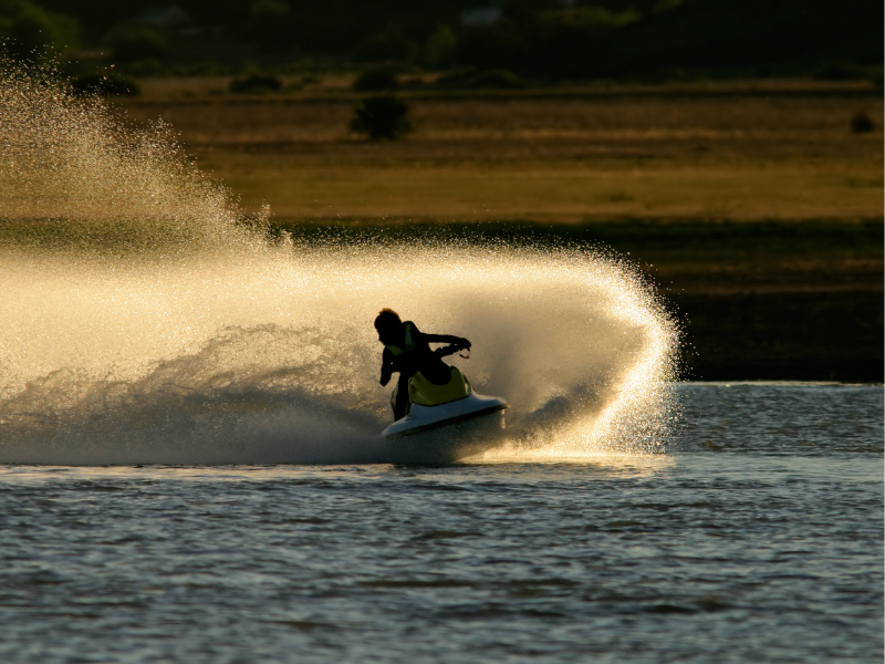 Pessoa pilotando um jet ski sobre a água, deixando um grande rastro dourado ao pôr do sol.