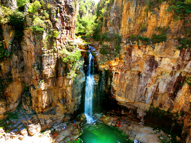 Alta cachoeira despencando em um cânion rochoso com penhascos alaranjados e vegetação verdejante.