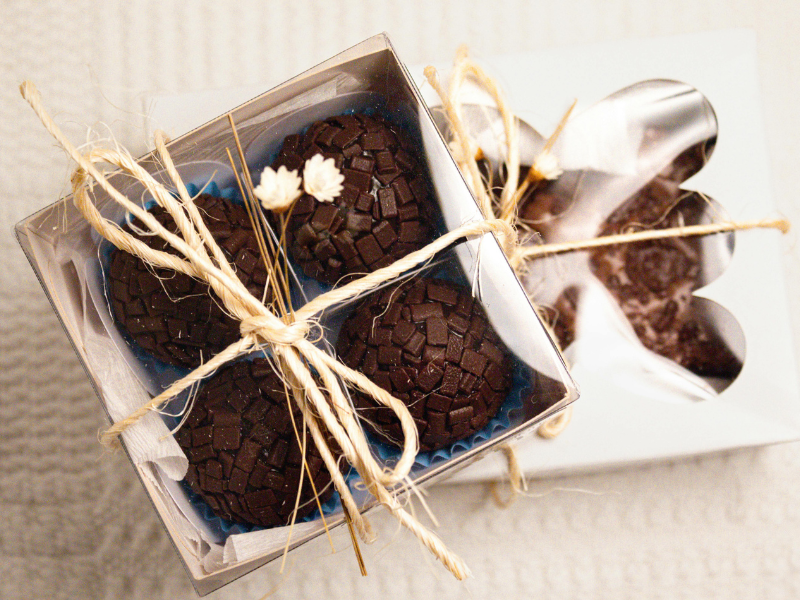 Duas caixas de presente amarradas, contendo bombons de chocolate, uma delas decorada com uma flor branca, sobre uma superfície clara.