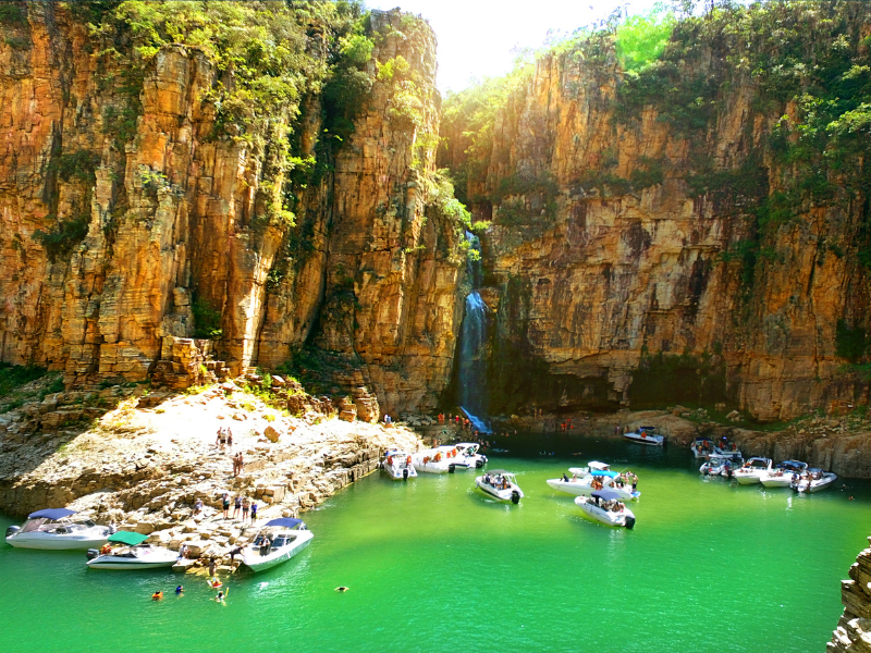 Lagoa turquesa com pequenos barcos ao lado de altas falésias alaranjadas e paredes rochosas verdejantes cobertas de floresta.