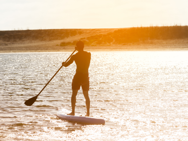 Pessoa praticando stand-up paddle em águas calmas ao pôr do sol, silhuetada contra a luz alaranjada.