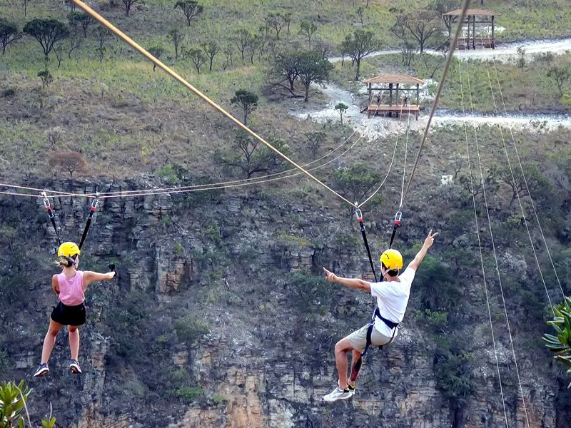 Duas pessoas com capacetes deslizando em tirolesa sobre um cânion rochoso acima de um rio.