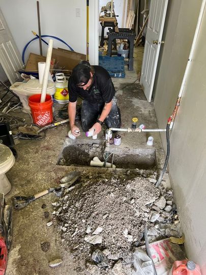 A professional kneeling on a concrete floor works on exposed pipes in an excavated trench amidst renovation debris.