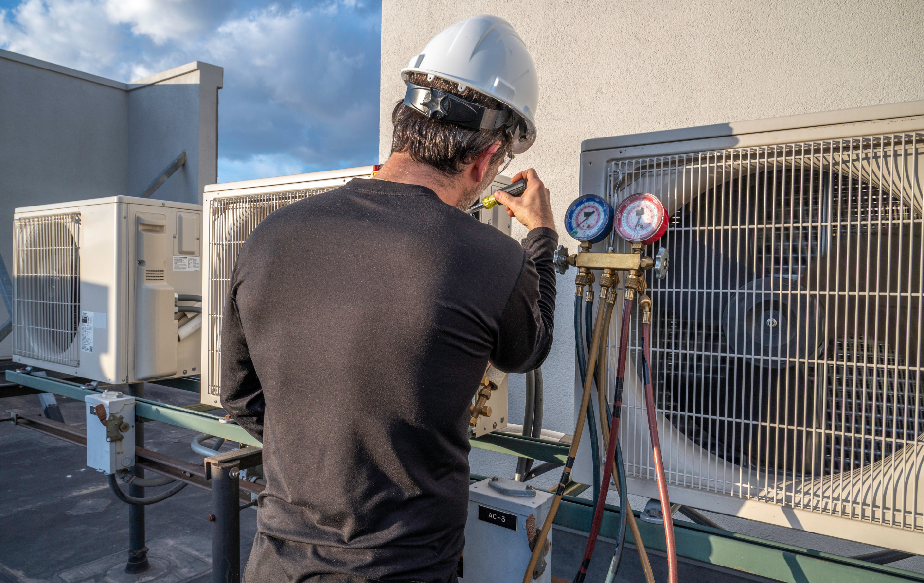 A technician in a white hard hat checks refrigerant pressure on an outdoor HVAC unit using a manifold gauge set.