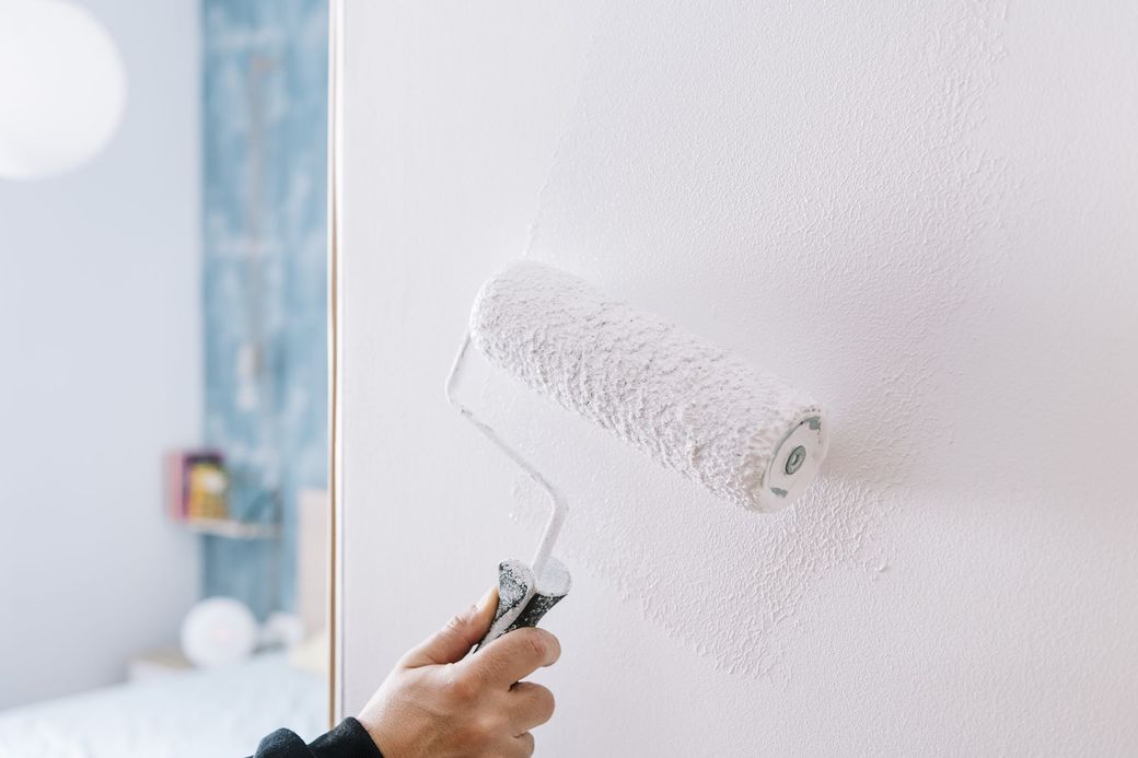 A hand uses a paint roller to apply white paint to a wall in a room.