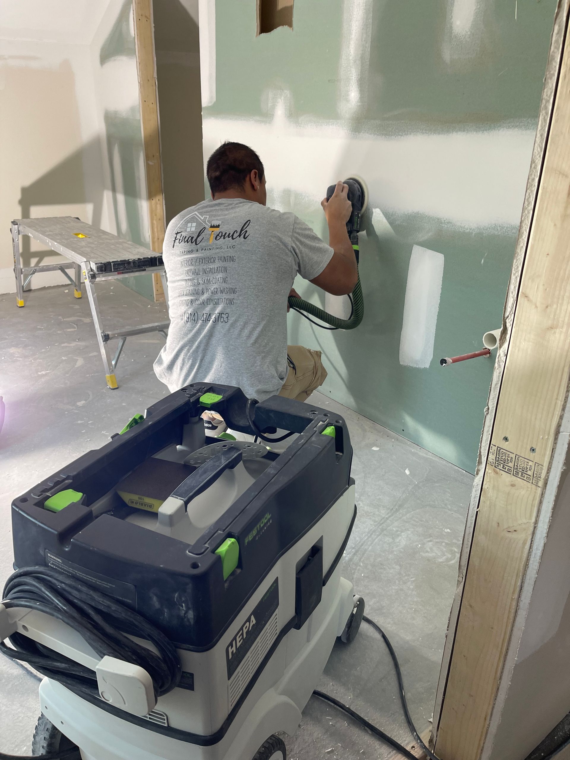 Worker sanding a drywall wall in a room with a floor vacuum and paint supplies