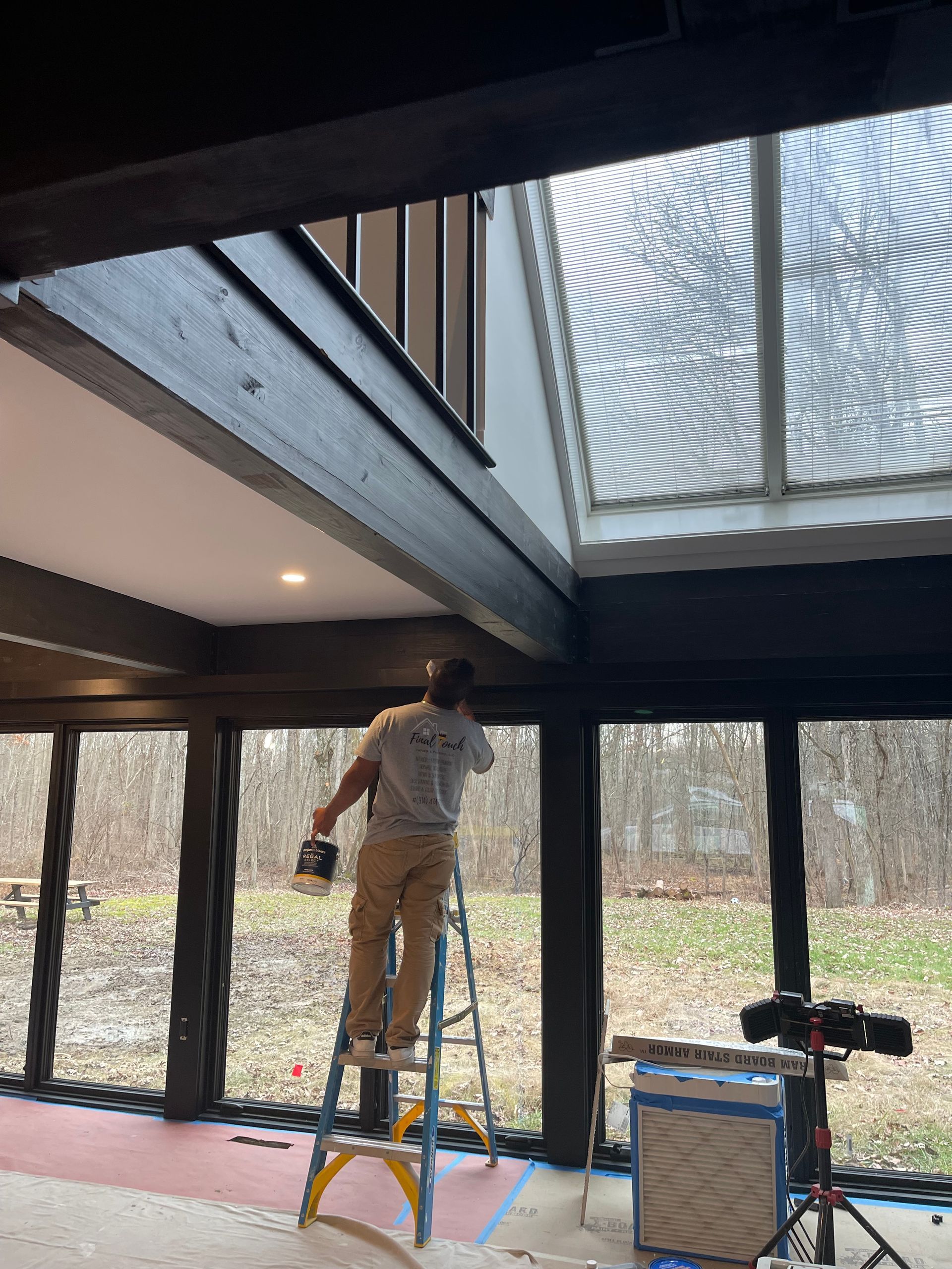 Person on a ladder painting a high ceiling in a modern room with large windows and skylight.