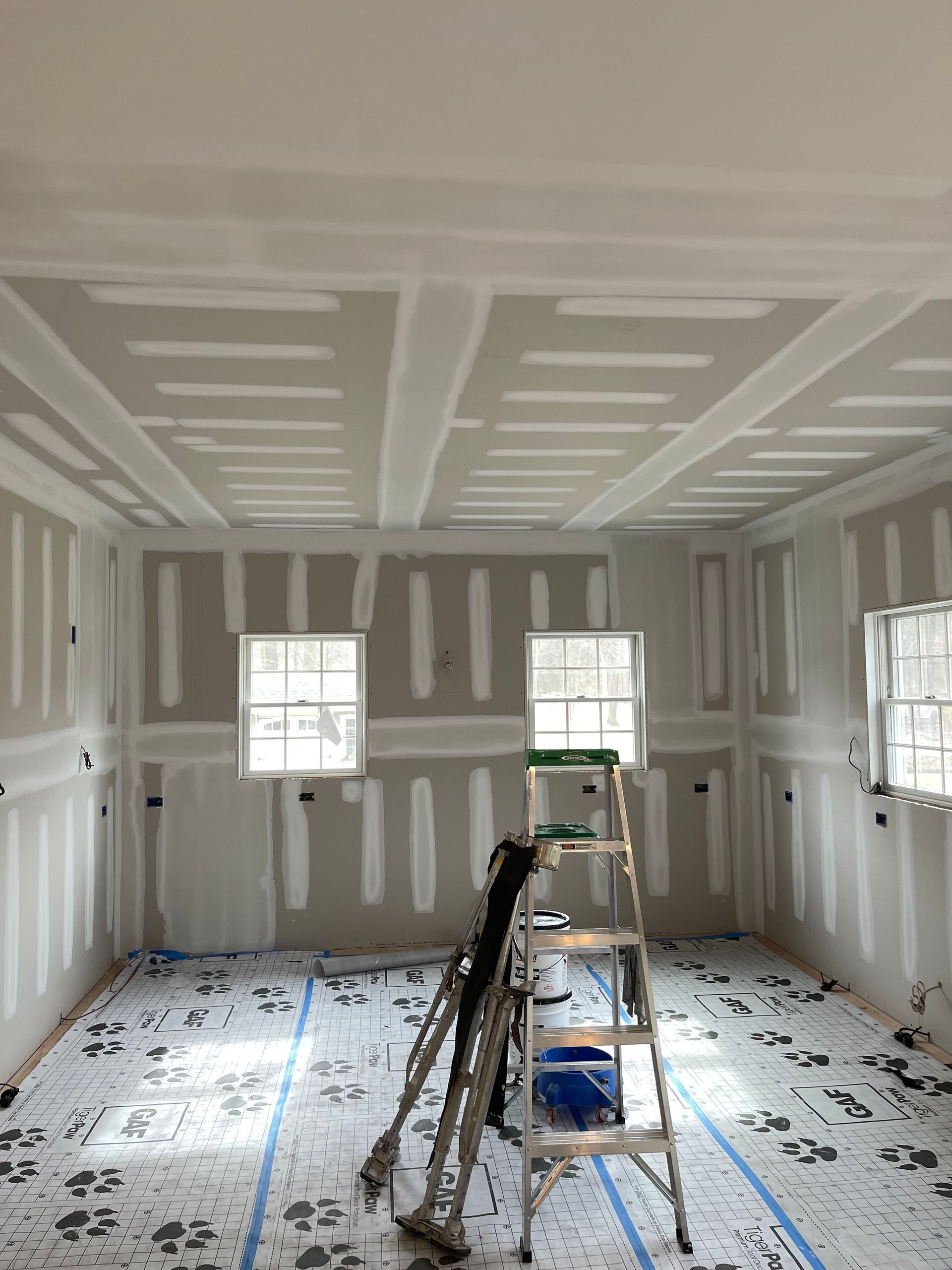 Unfinished room with drywall, windows, and a ladder on a taped floor during renovation.