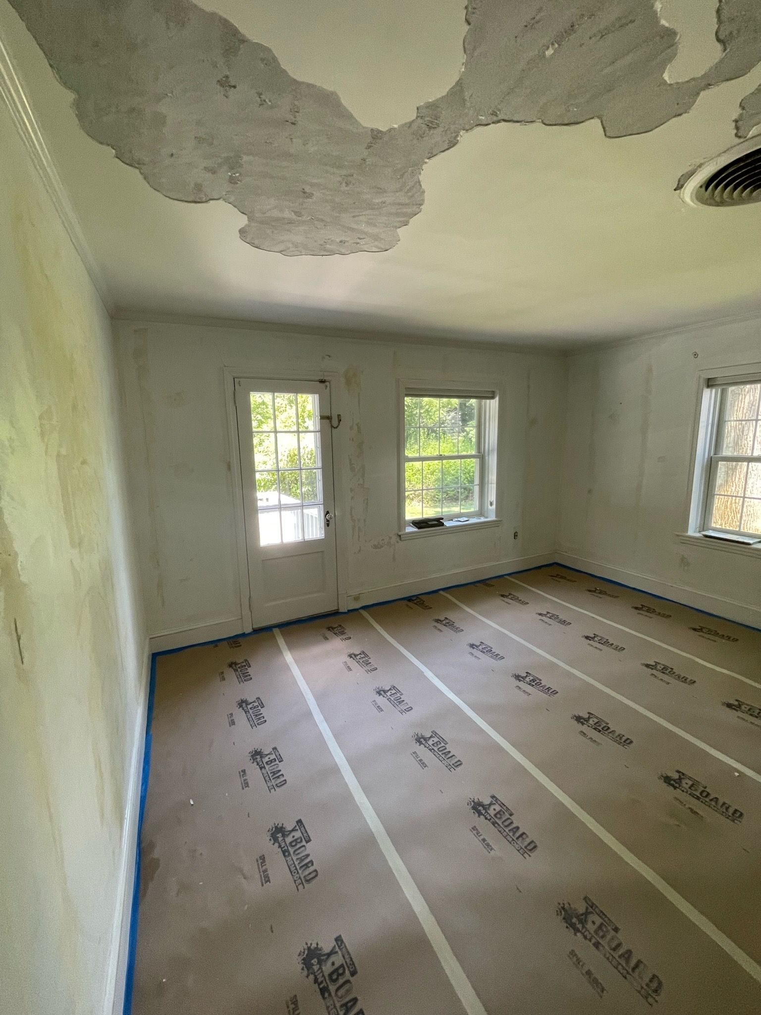 Empty room with peeling ceiling paint, three windows, and protective floor covering during renovation
