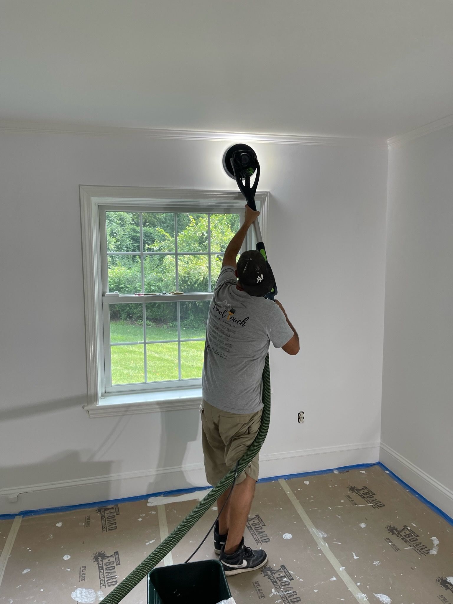 Person painting a white room ceiling near a window, with floor covered and blue tape along the baseboards