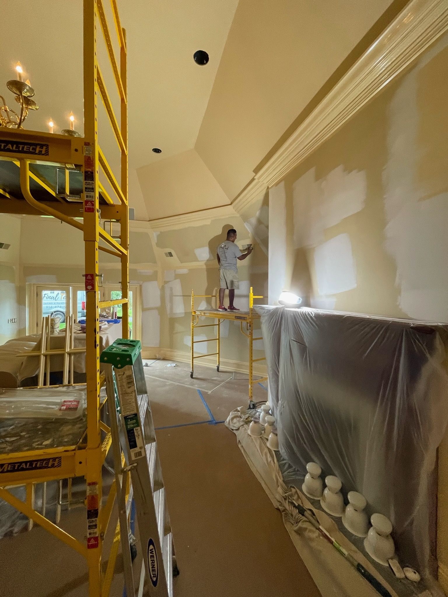 Worker painting a high wall on scaffolding in a room under renovation, with tarps and supplies below.