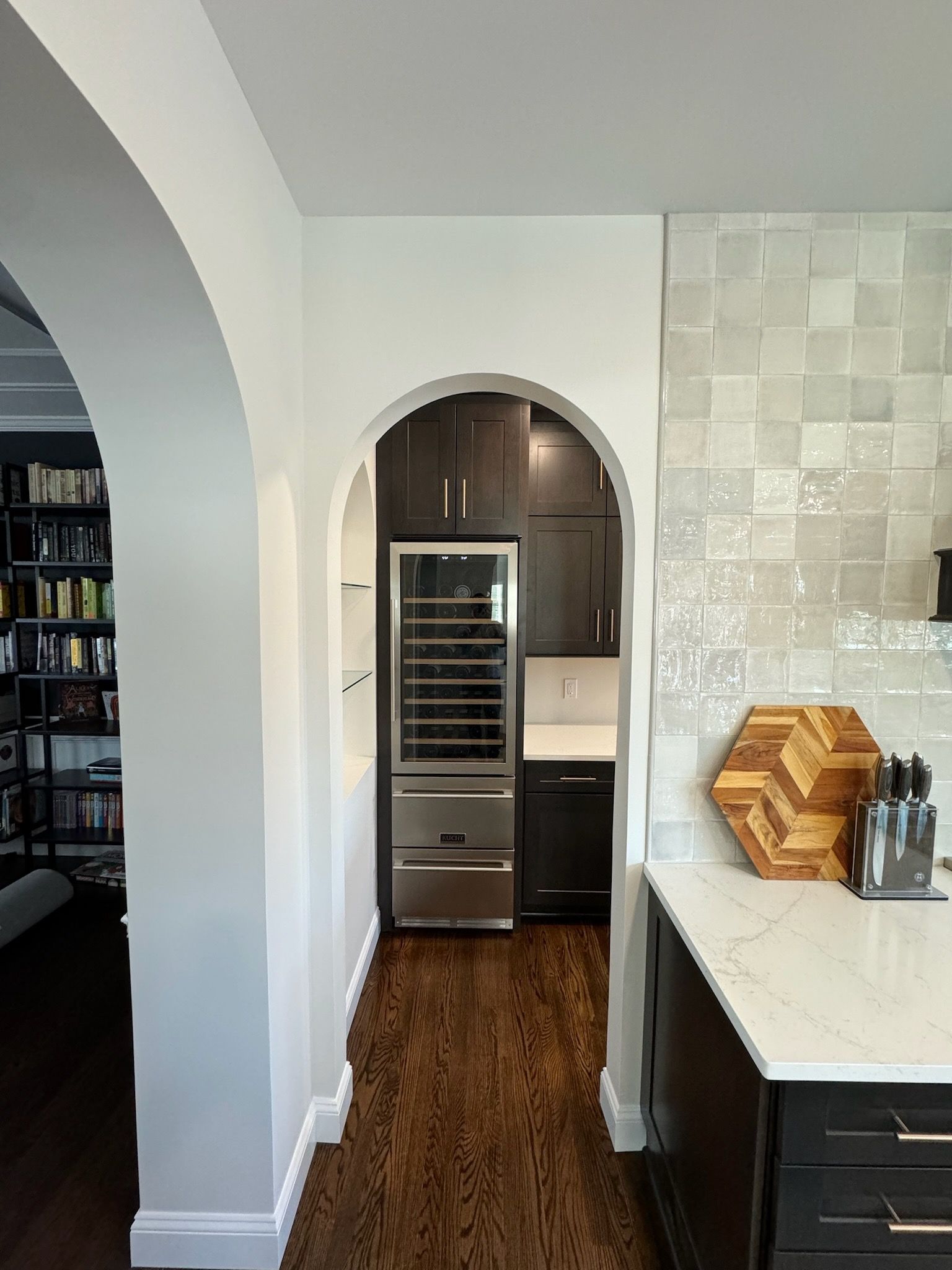 Kitchen view through arched doorway to a wine fridge, dark cabinets, and white countertop with stacked bowls.