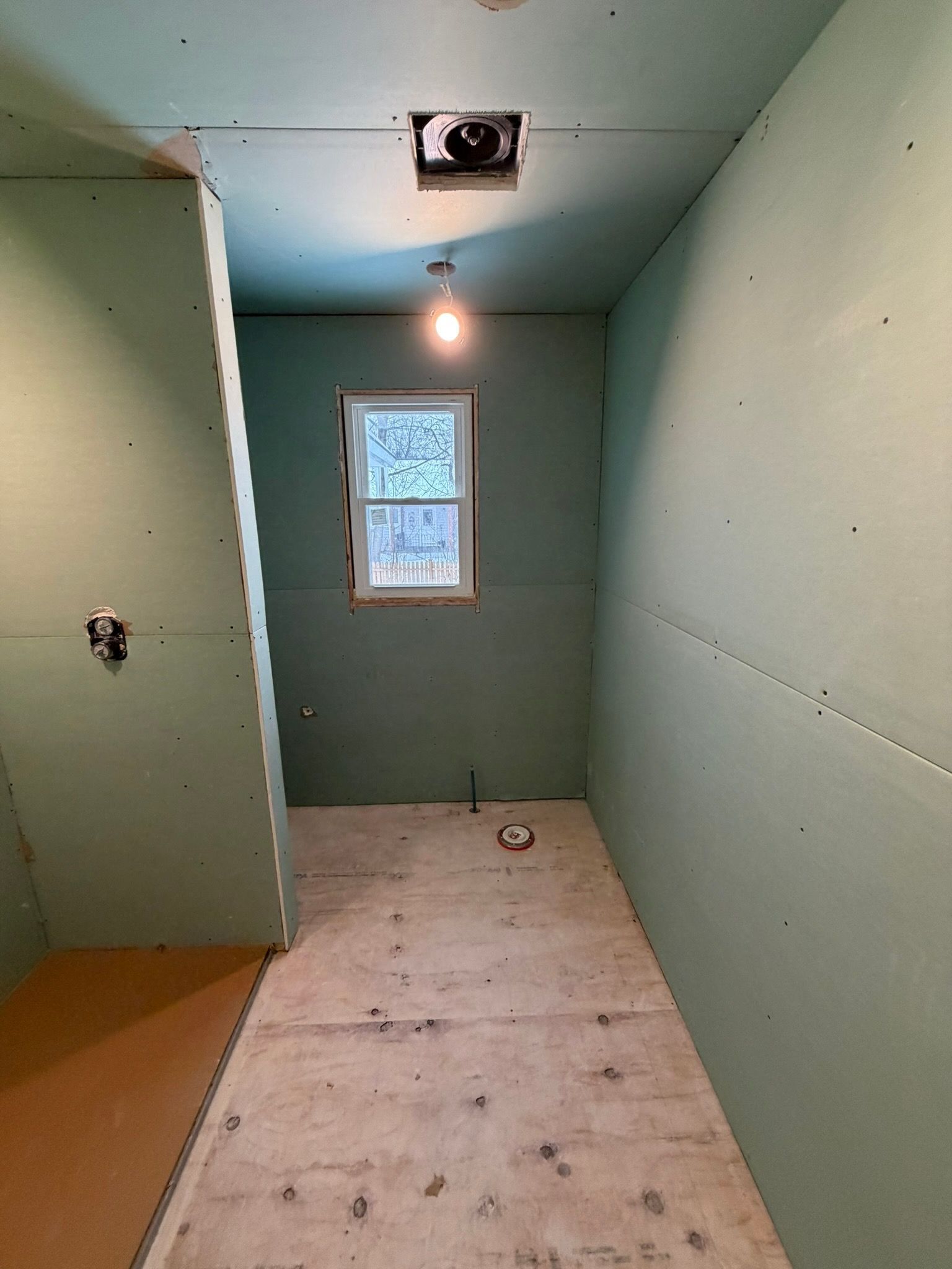 Partially finished bathroom with green drywall, window, ceiling light, and toilet rough-in hole on the floor