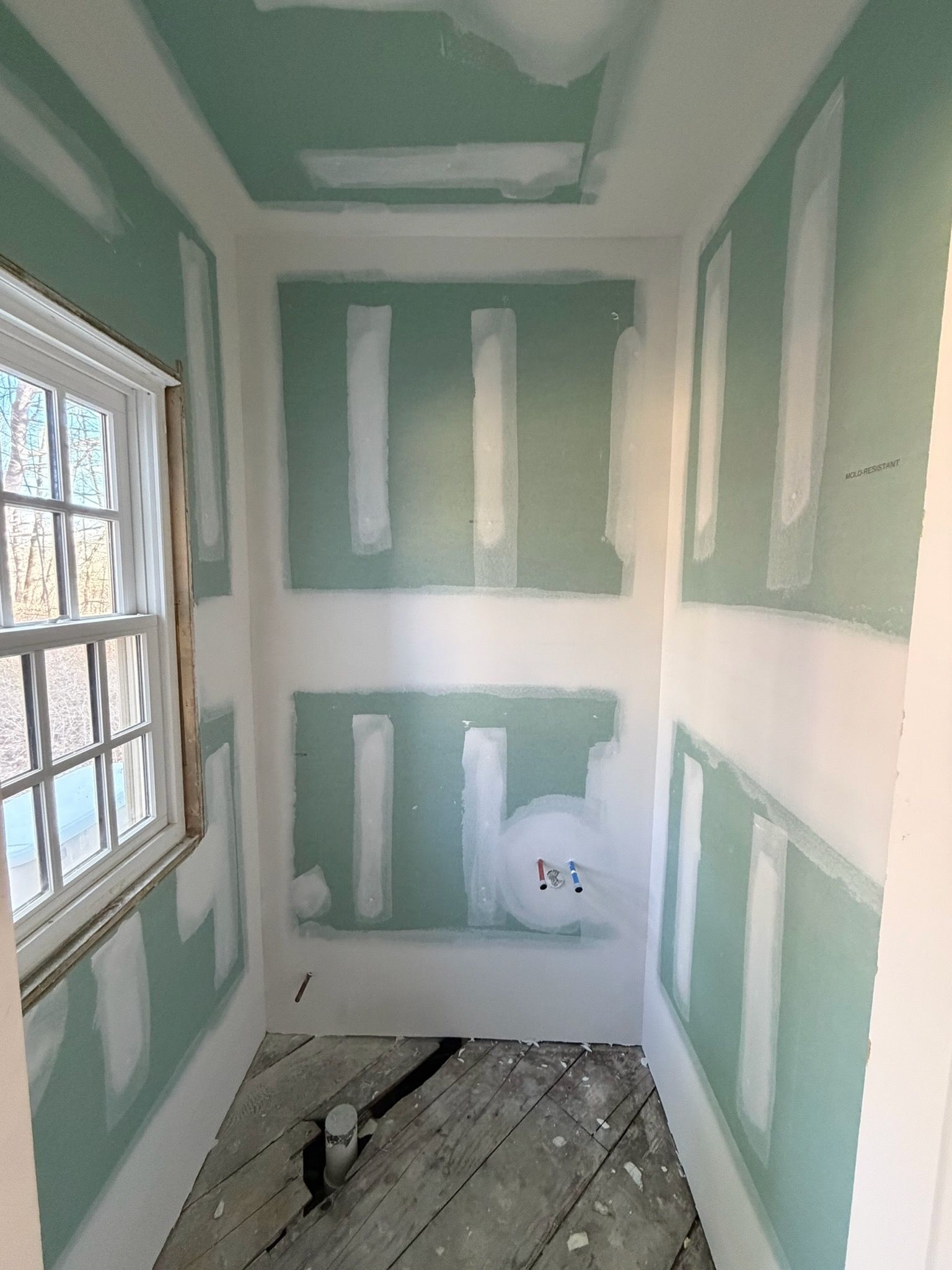 Narrow unfinished room with green drywall patches, a window, and exposed floorboards.
