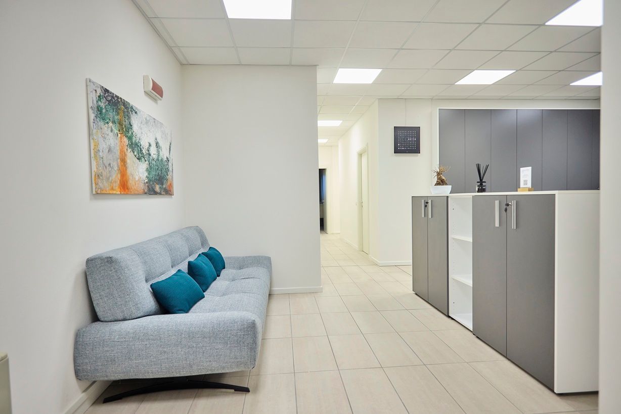 Una sala d'attesa con un divano grigio chiaro con cuscini color verde acqua, pareti bianche decorate con quadri