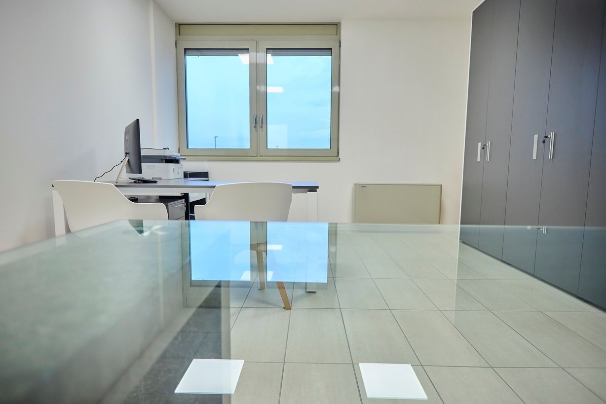 Un ufficio caratterizzato da una scrivania in vetro in primo piano, sedie bianche, una postazione computer