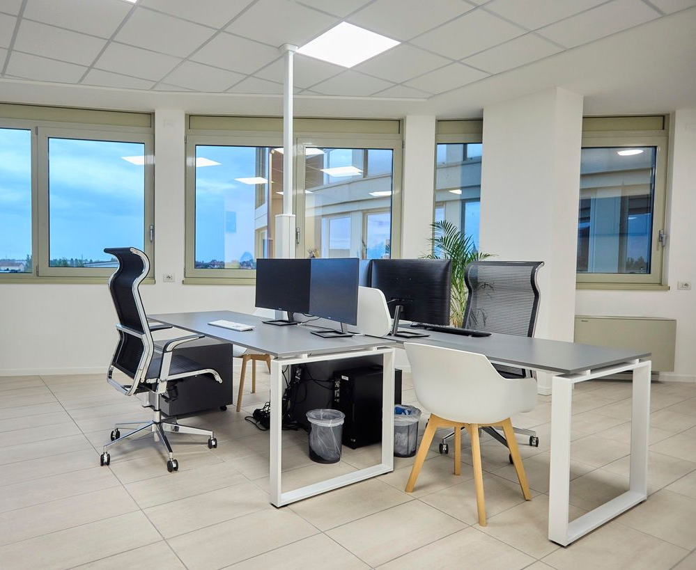 Moderno ufficio con due scrivanie, monitor e sedie vicino a grandi finestre, illuminato da luminose luci a soffitto.