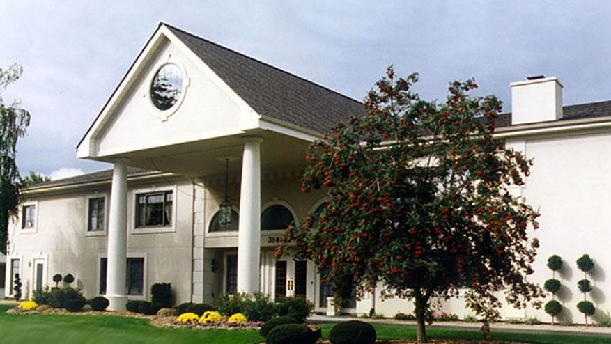 White building with columns, circular window, and landscaped grounds on a sunny day