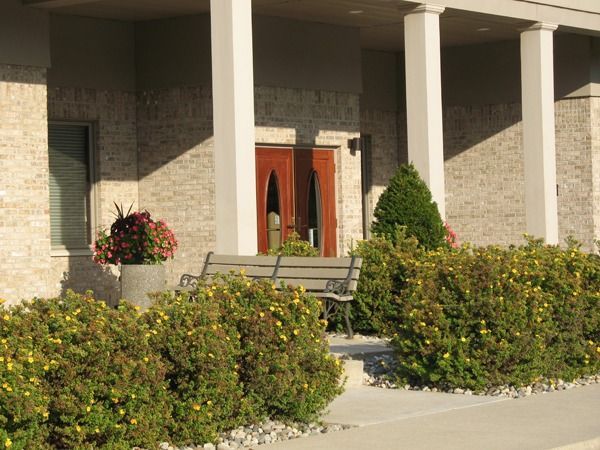 Front entrance of a brick house with white columns, potted flowers, and trimmed shrubs