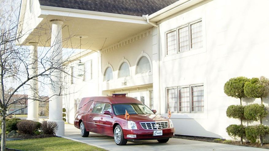 Red minivan parked in a driveway beside a white house with columns and trimmed bushes.