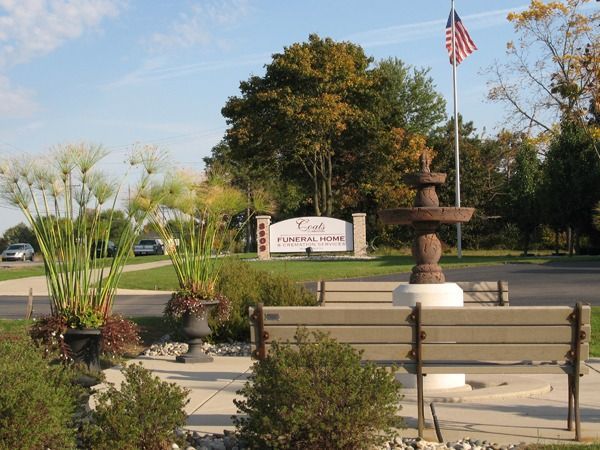 Park entrance sign with fountain, benches, flowers, and an American flag on a sunny day