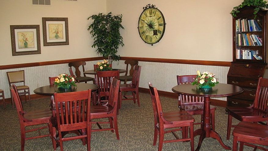 Small dining room with wooden tables and chairs, wall clock, framed art, plant, and bookshelf