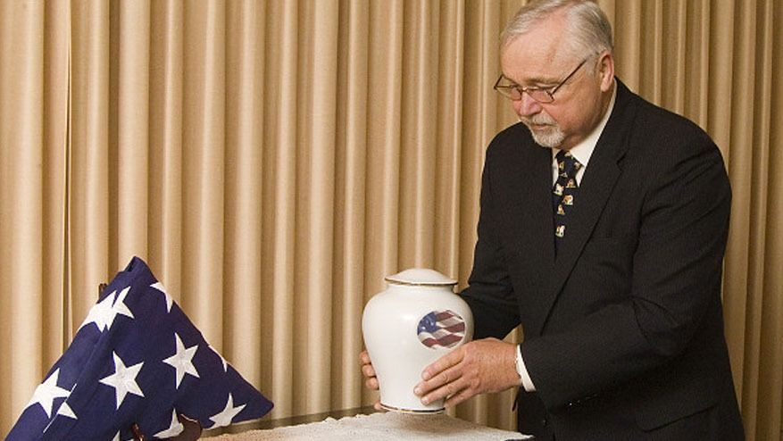 Man in a black suit placing a folded U.S. flag beside a white funeral urn on a table