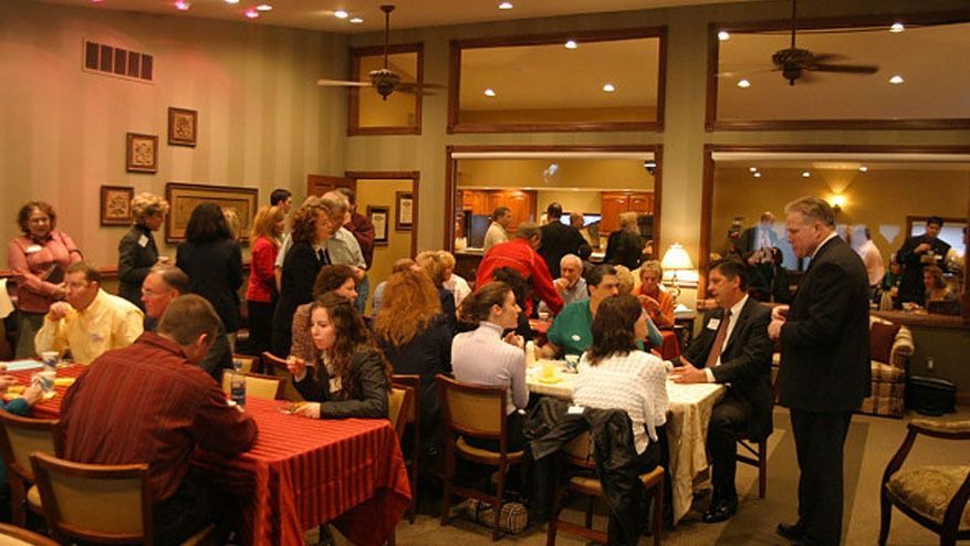 Crowded restaurant gathering with people seated at tables and a speaker addressing the room