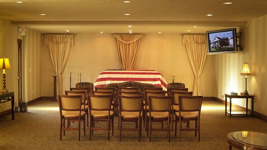 Small memorial room with a casket draped in a red-and-white flag, surrounded by chairs and warm lamps