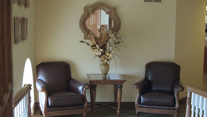Two dark brown armchairs flanking a small table with flowers beneath a decorative wall mirror in a hallway.