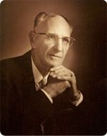 Sepia portrait of a man in glasses, resting his chin on clasped hands, in a dark suit and tie