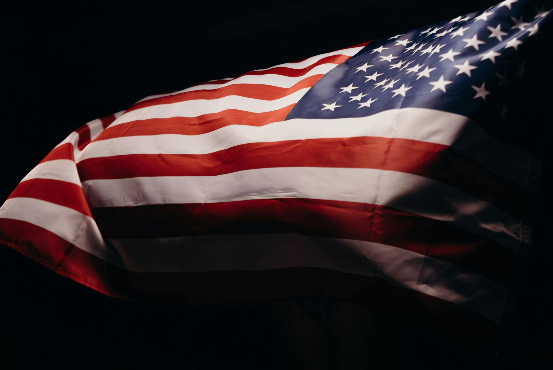 Waving American flag against a dark background, red, white, and blue stripes lit dramatically