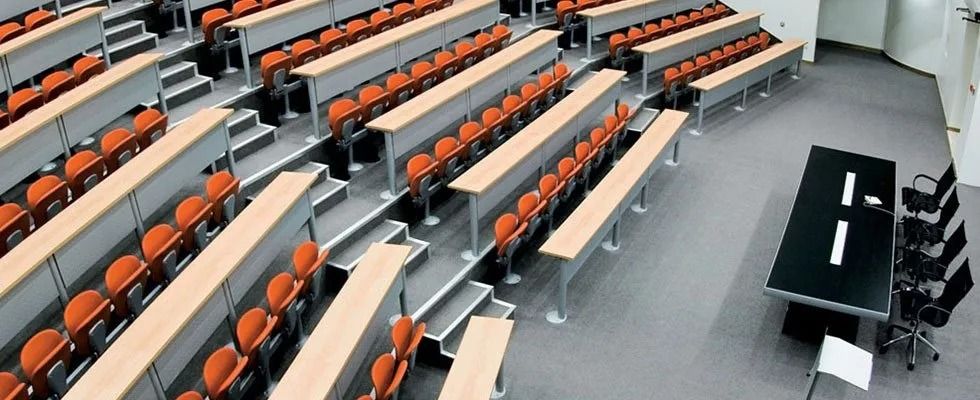 Aula magna vuota con sedili arancioni, lunghi banchi e un tavolo nero per le presentazioni davanti