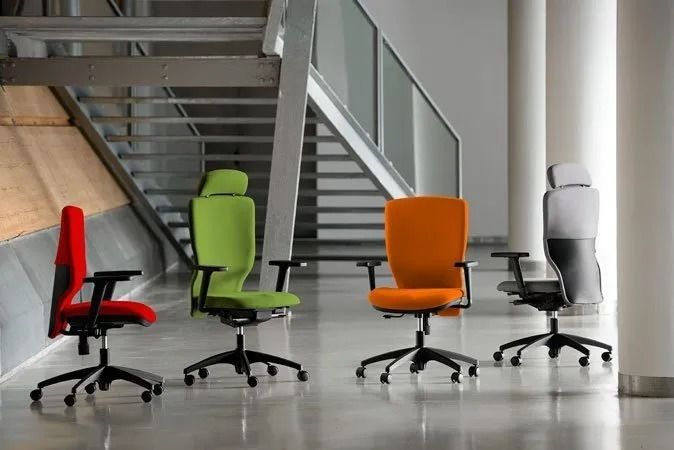 Quattro moderne sedie da ufficio, rosse, verdi, arancioni e bianche, in un luminoso atrio con scale.