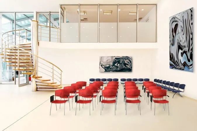 Moderna sala conferenze con sedie rosse e blu, una scala a chiocciola e opere d’arte astratte alle pareti