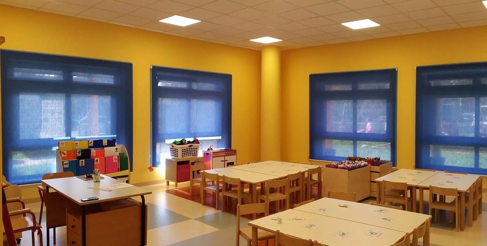 Aula luminosa con pareti gialle, finestre blu e tavoli disposti per le attività.