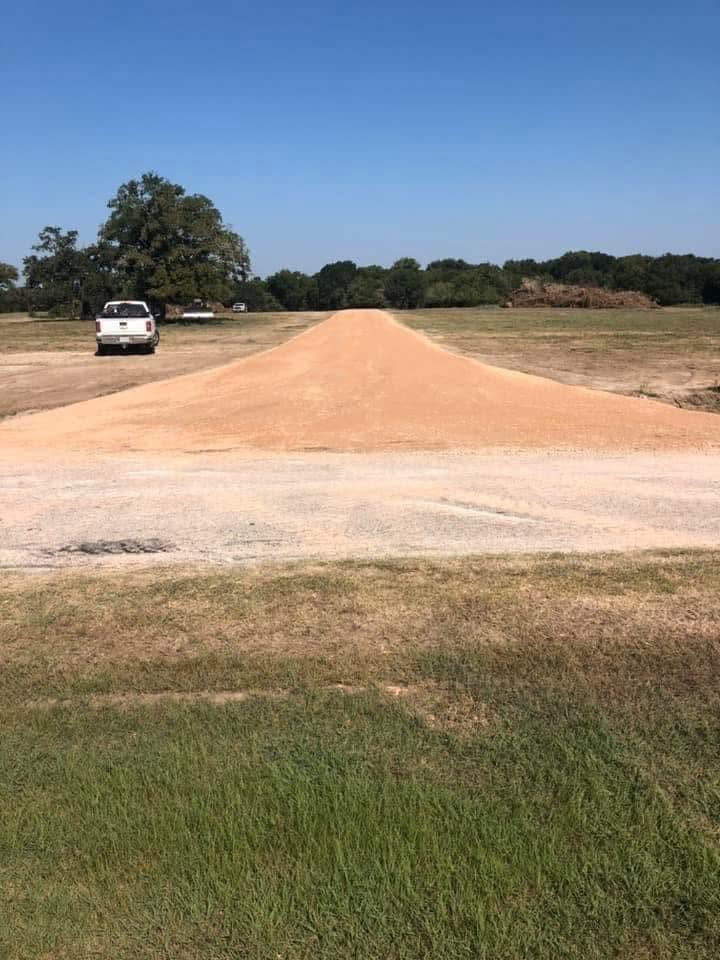 Dirt road under construction with a white SUV parked on the left and grassy fields under a clear blue sky